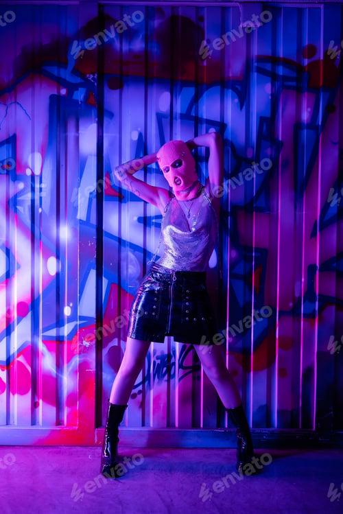 Preview: full length of woman in glamour outfit and balaclava posing with hands behind head near graffiti in