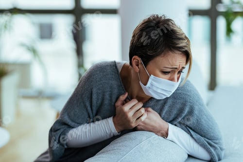 Preview: Sick woman wearing face mask while coughing at home.