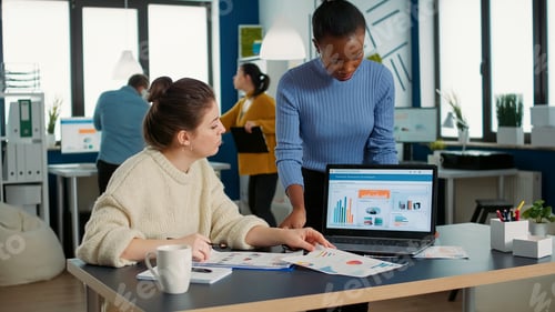 Preview: African american woman holding laptop asking colleague looking at clipboard with charts for advice