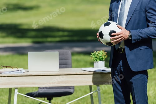 Preview: cropped view of businessman holding soccer ball while standing in park near table with laptop and