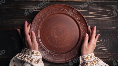 Preview: Female hands with empty ukrainian clay plate from wooden table. Woman in traditional embroidered