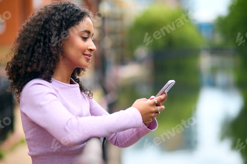 Preview: Young Woman Standing By River In Urban Area Using Mobile Phone For Messaging Or Social Media