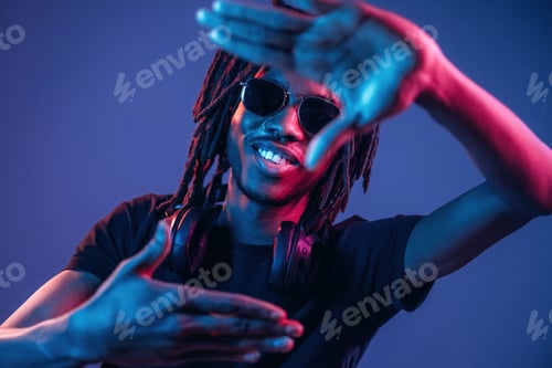 Preview: Gesture of capturing the photo. African American guy is studio with neon lighting