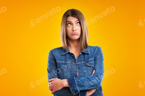 Vista previa: Close up of woman in denim jacket thinking and pointing over yellow background