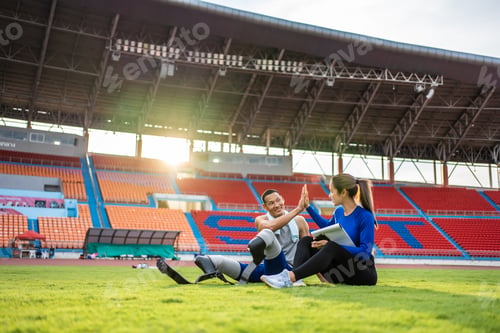 Preview: Asian para-athlete with prosthetic blades and trainer sit in stadium.