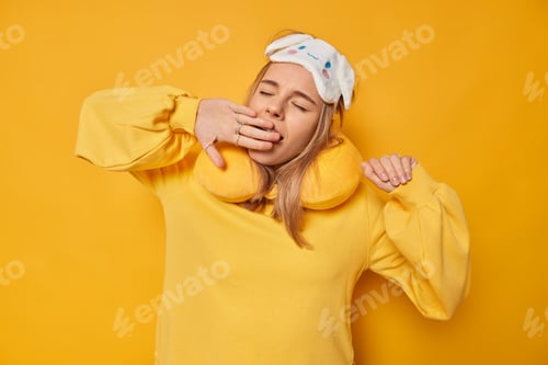 Preview: Tired young woman wants to sleep covers opened mouth as yawns wears sleepmask neckpillow dressed in