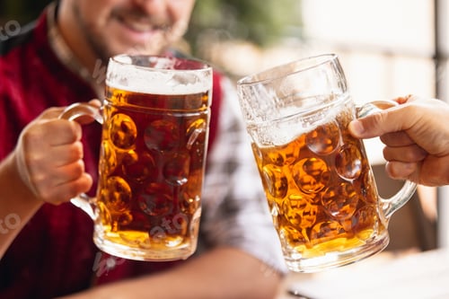 Preview: Close-up two mugs, glasses of beer in male hands. Friends meeting at bar, cafe or pub. Traditions