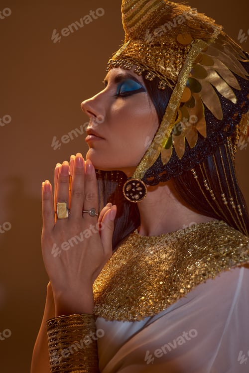 Preview: Side view of woman in Egyptian attire and headdress doing praying hands gesture on brown background