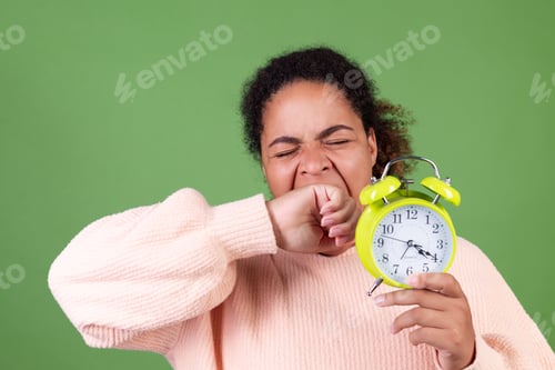 Preview: Beautiful african american woman on green background with alarm clock sleepy tired exhausted yawning