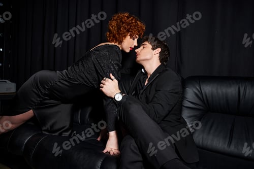 Preview: enticing boyfriend and girlfriend in elegant clothes hugging lovingly on black sofa, sexy couple