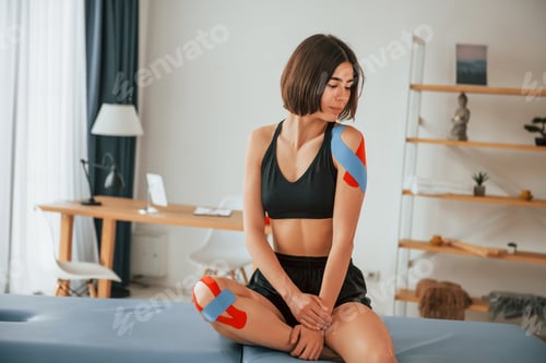 Preview: Sitting on the table. Woman with kinesio tape is in the clinic indoors