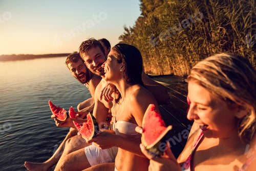 Preview: Friends sitting on the edge of a pier enjoying on a summer day at the lake eating watermelon.