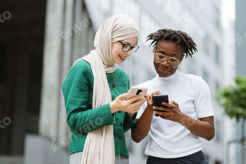 Preview: Woman in hijab withsmartphone and african female with phone standing together near office building
