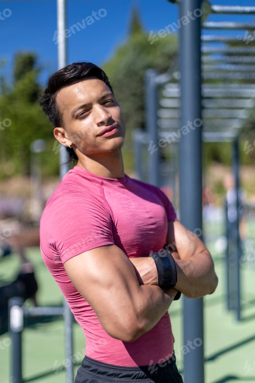 Preview: Confident young man in pink shirt posing with arms crossed at outdoor gym.