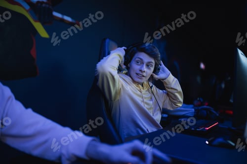 Preview: Young gamer in headphones talking to blurred friend in cyber club with lighting