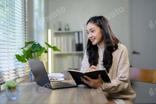 Preview: Young asian businesswoman is taking notes in her notebook while working from her home office, using