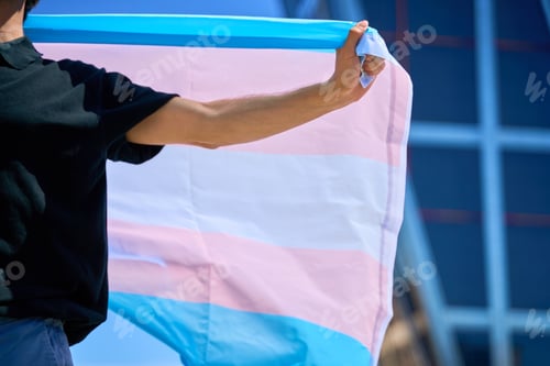 Preview: A man holding a transgender flag in front of a building. The flag is large and colorful.