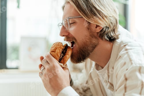 Preview: Young freelancer man with beard and glasses enjoying eating burger at home office