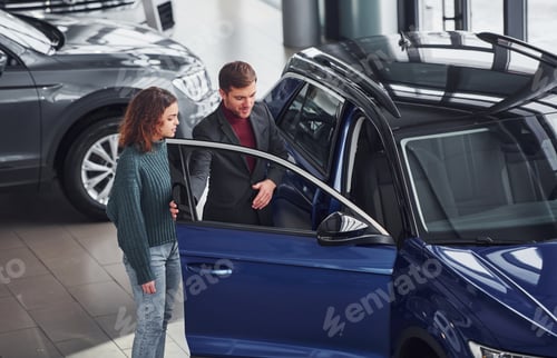 Preview: Professional salesman assisting young girl by choosing new modern automobile indoors