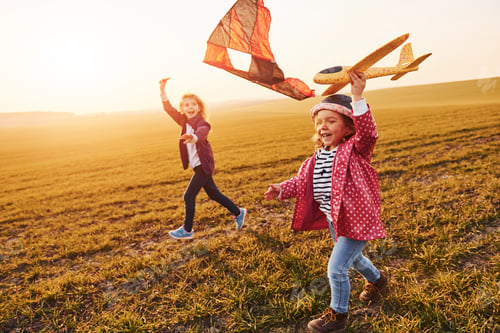 Preview: Two little girls friends have fun together with kite and toy plane on the field at sunny daytime