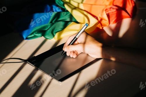 Preview: Designer works on graphic tablet indoors, rainbow flag symbol LGBT community on table. Close-up of