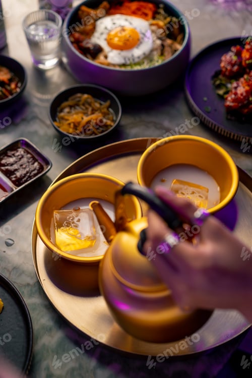 Preview: girl pouring a traditional Korean low-alcohol drink makoli from a golden teapot into a cup with ice
