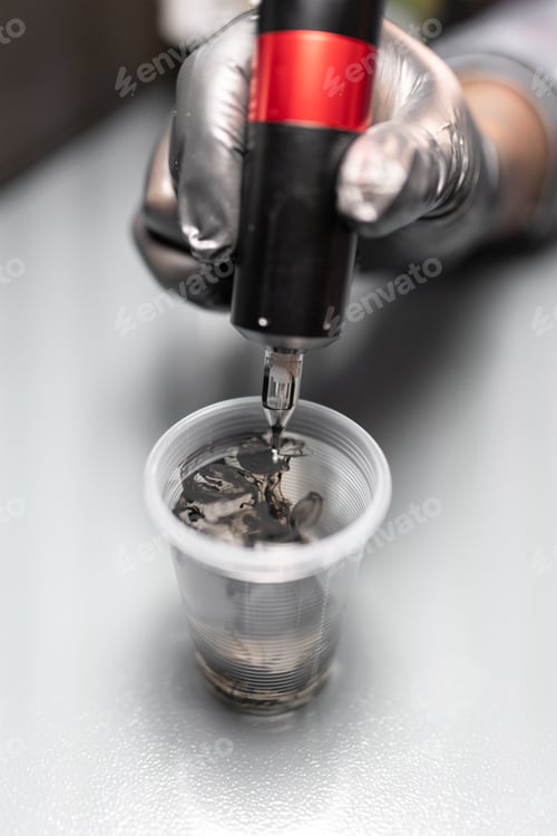 Preview: Close up of a tattoo artist is pouring ink into a plastic glass to clean the needle