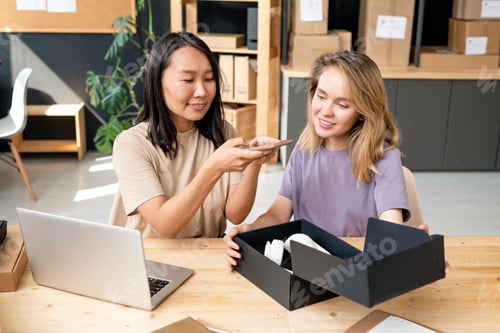 Preview: Happy young Asian woman taking photo of headphones with her colleague near by
