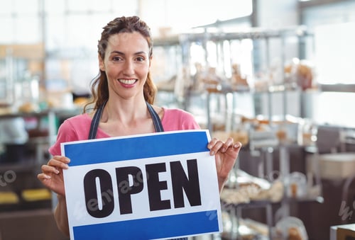 Preview: Smiling woman baker holding OPEN sign inside bakery, with metal racks and pastries