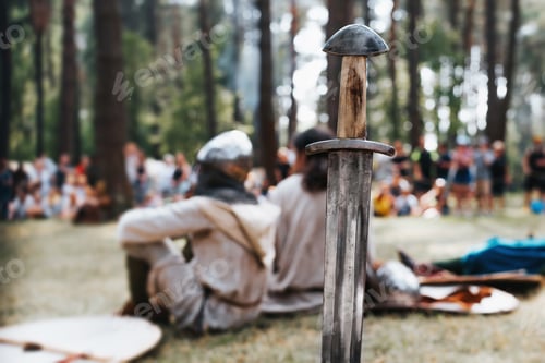 Preview: Medieval old steel sword against the background of knights sitting outdoors.