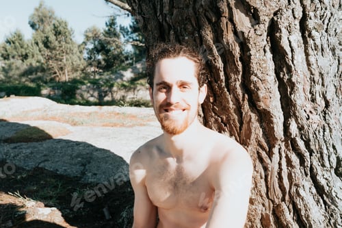 Preview: Young man with a beard next to a tree shirtless smiling to camera during a sunny day