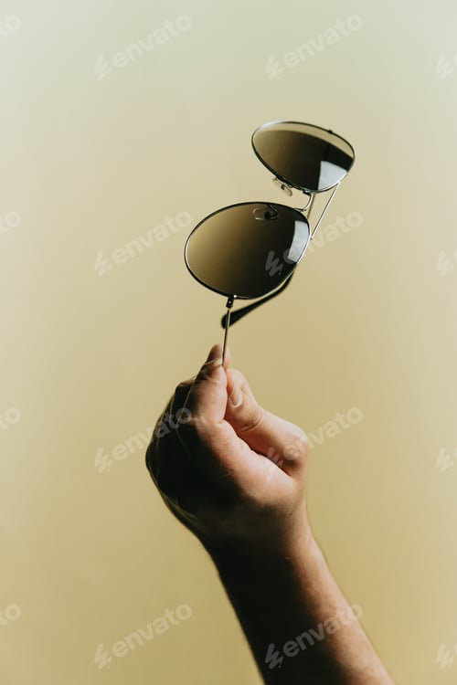 Preview: Hand grabbing a pair of dark sunglasses over a pastel yellow background, style and minimal