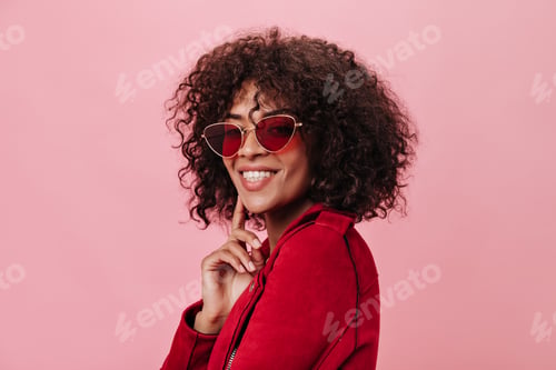 Preview: Charming girl in red jacket and bright glasses is smiling on pink background. Curly woman in stylis
