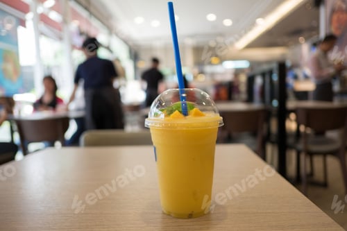 Preview: A glass of mango smoothie on a wooden table in a restaurant with blurred people in background