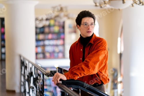Preview: portrait of a young guy in glasses standing with a book in his hands in library