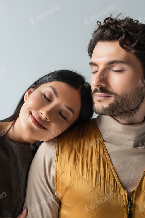 Preview: smiling asian woman with closed eyes leaning on stylish brunette man isolated on grey