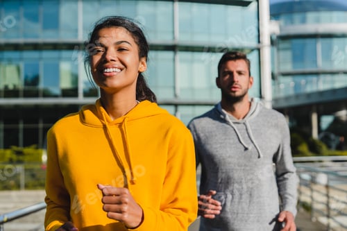 Preview: Smiling fit Afro girl in the foreground jogging with handsome caucasian male runner in urban area