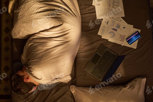 Preview: Young Man Sleeping in Bed Surrounded by Laptop and Documents
