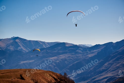 Preview: Floating serenely in the clear sky, paragliders soar above the rugged brown mountain peaks.