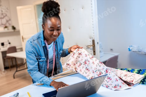 Preview: Female fashion tailor working on dress design in her workshop using laptop.