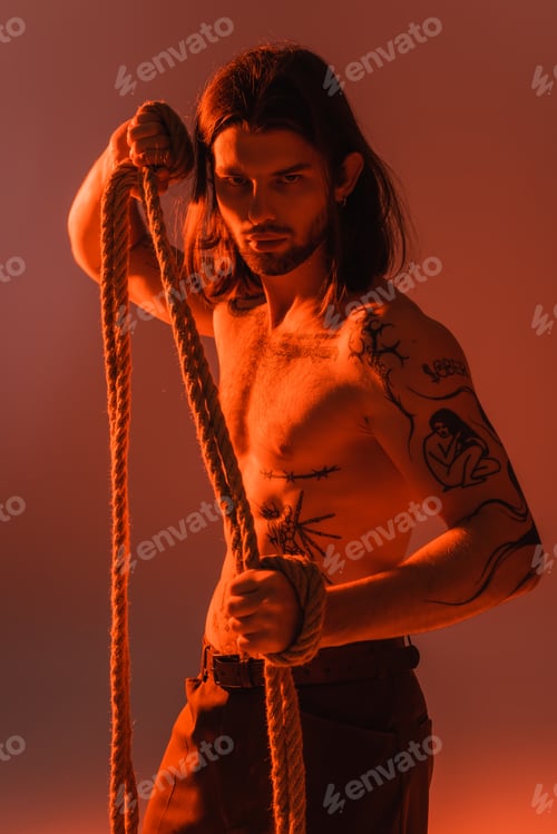 Preview: Long haired and tattooed model holding rope and looking at camera on red background