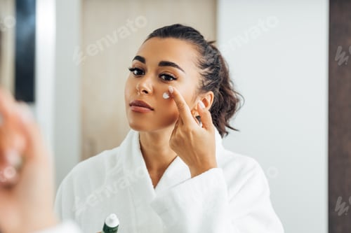 Preview: Beautiful woman in bathrobe applying anti-aging cream on her cheek