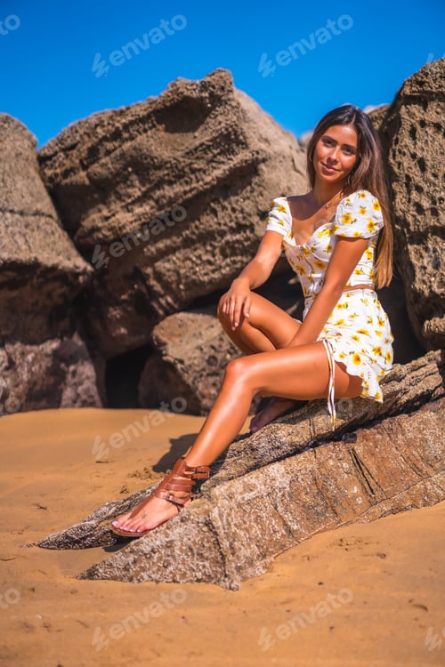 Preview: Portrait of a brunette girl in a white dress on the beach enjoying the summer, sitting on some rocks