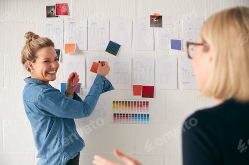 Preview: Female Business Owner With Intern Looking At Designs On Wall In Start Up Fashion Business