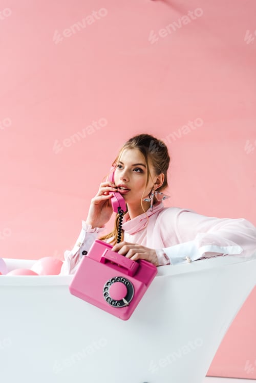 Preview: pretty girl talking on retro phone while lying in bathtub on pink