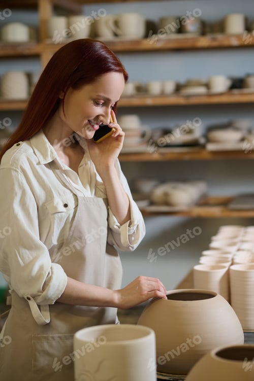Preview: Young woman talking on the phone while standing in a pottery workshop