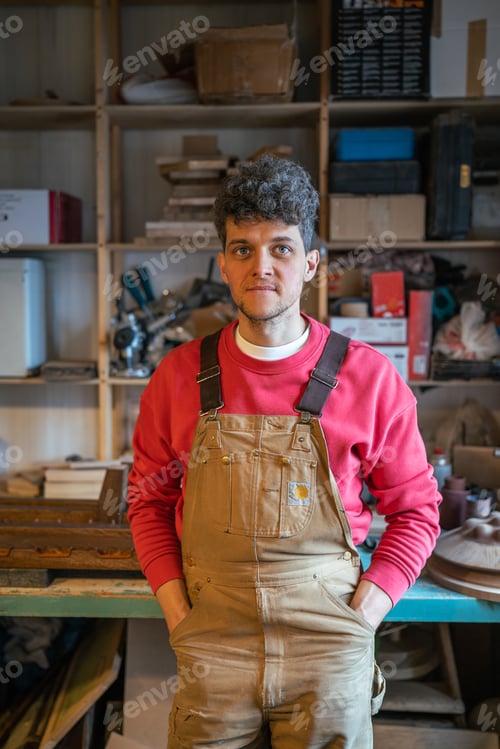 Preview: Smiling man carpenter resting in workshop looking at camera putting hands in pockets leans on table