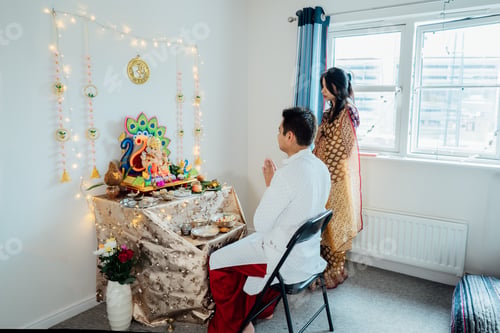 Preview: Indian couple dressed in traditional wear engages in worship at home altar, adorned for god Ganesh