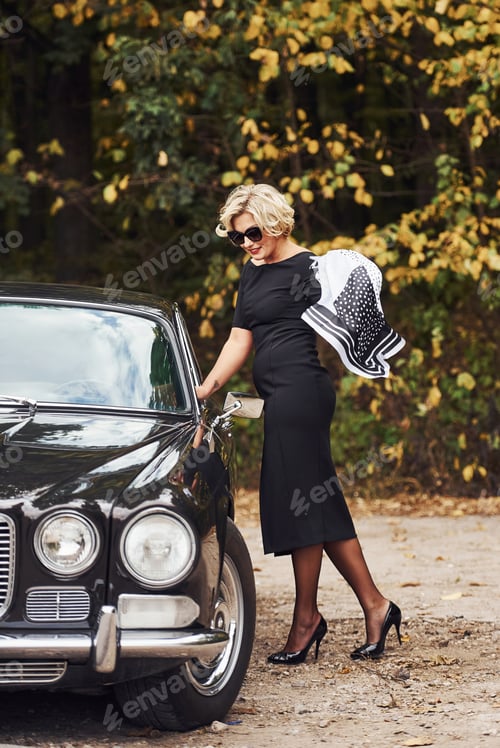 Preview: Blonde woman in sunglasses and in black dress near old vintage classic car