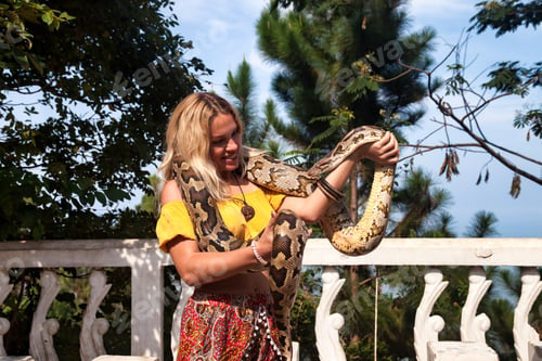 Preview: Happy woman tourist in yellow shirt holding snake python on shoulders and hands